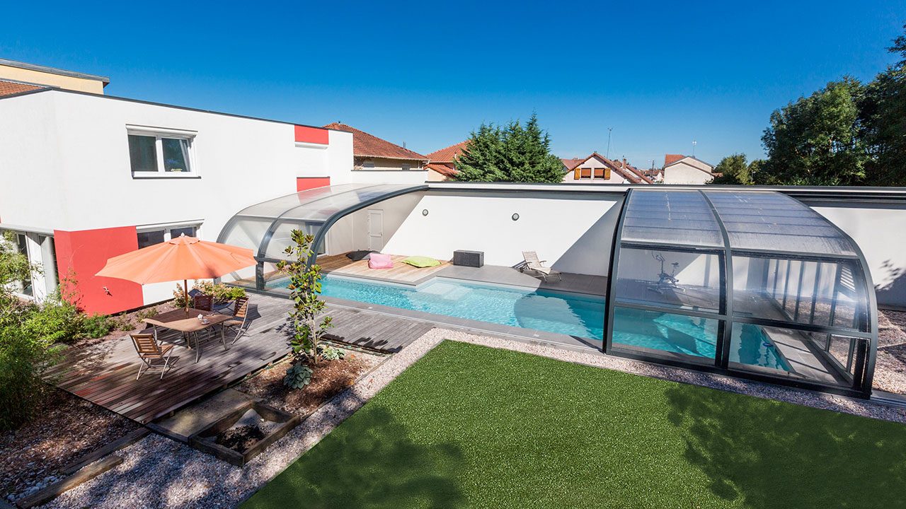 Piscine couverte par un abri bas vitré, terrasse en bois et gazon